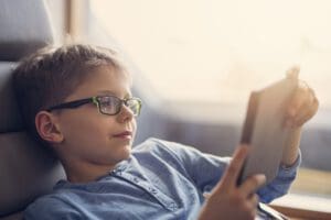 Happy boy aged 8 reading an e-book using e-reader. The boy is sitting comfortably in armchair