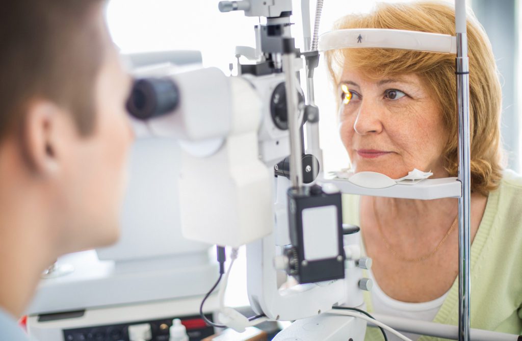 Senior caucasian woman having her eyes examined at the optician. Her head is placed in phoropter apparatus while middle aged male doctor is examining her retina. The woman has mid length yellow brown hair and wearing light breen blouse.