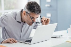 Businessman working at office desk, he is staring at the laptop screen close up and holding his glasses, workplace vision problems